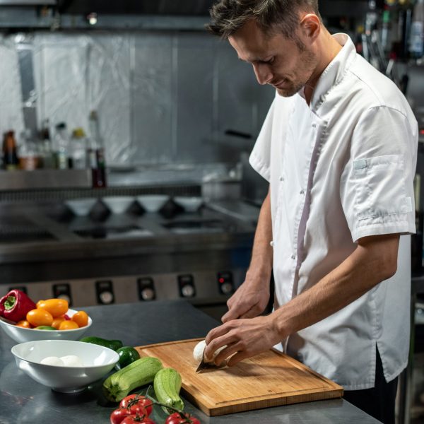 Young professional chef of modern restaurant in white uniform standing by large table in the kitchen and cutting onion and other vegetables